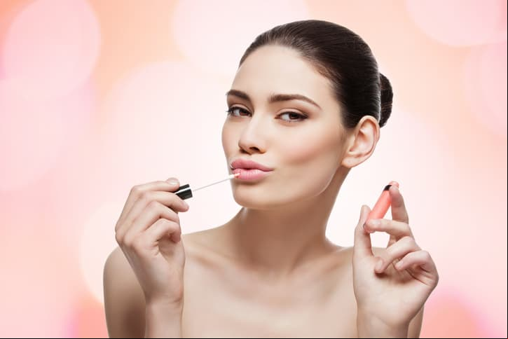 Beautiful young woman applying coral color lipgloss on lips. Isolated over white background, Copy space.