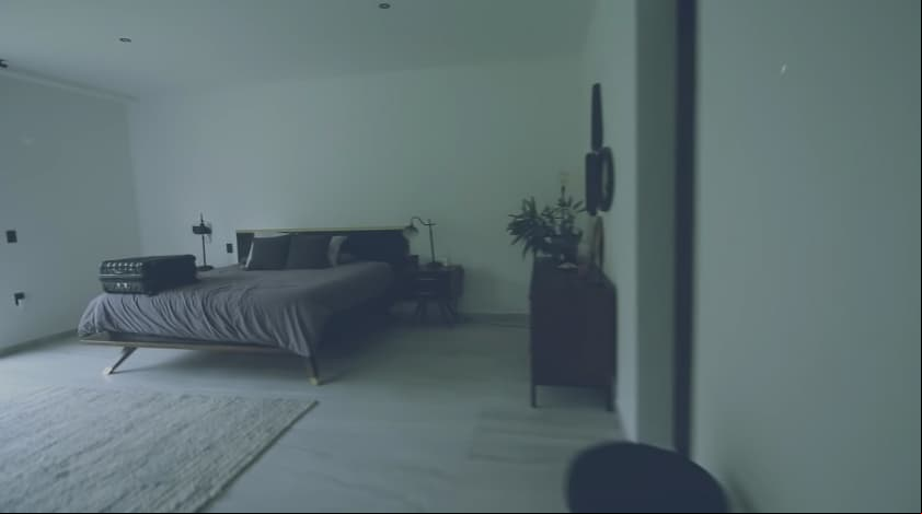 La habitación cuenta con una amplia cama con un buró, una lámpara y una vela, todo en tonalidades oscuras, además de contar con un gran espejo.