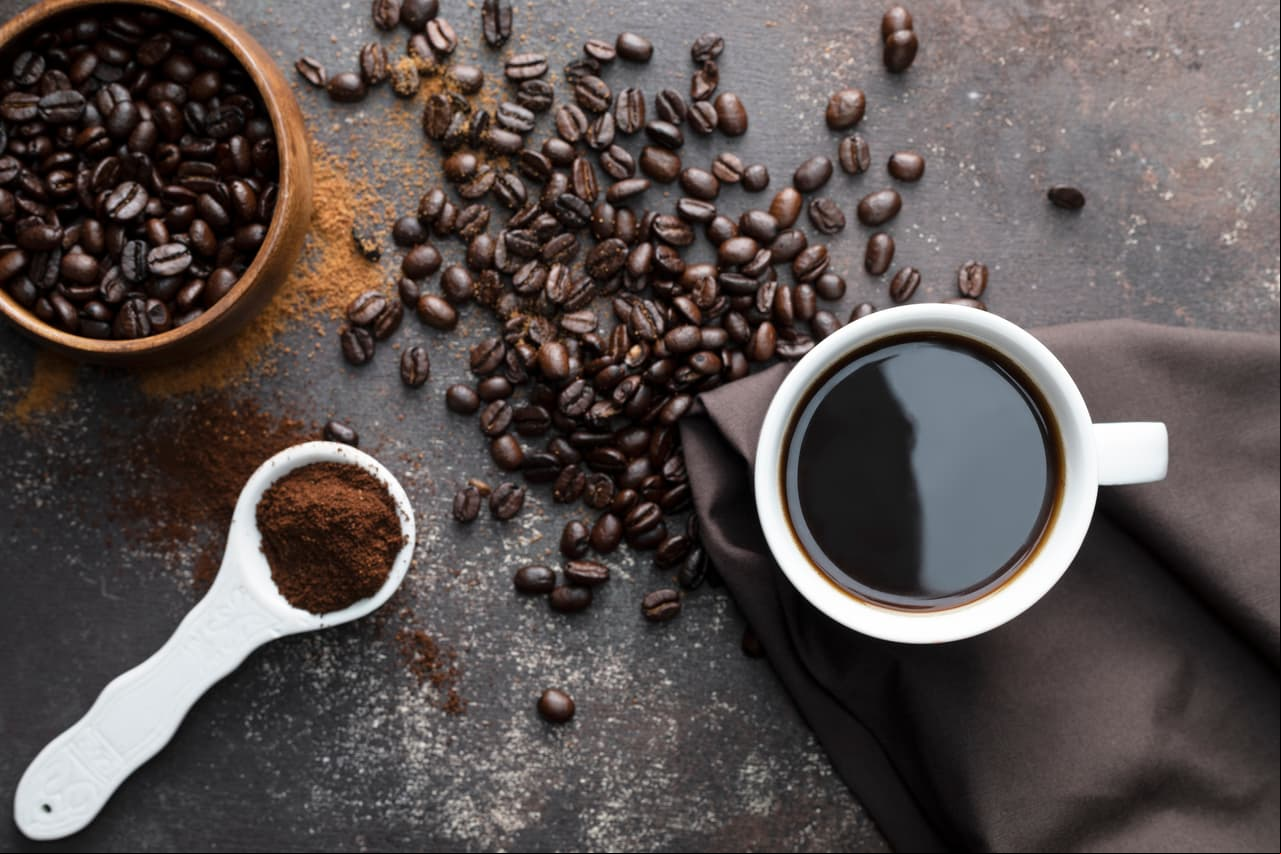 Direct above view of roasted coffee beans, white coffee cup with black coffee, dark brown colored napkin with one white spoon full with blended coffee on dark brown background.
